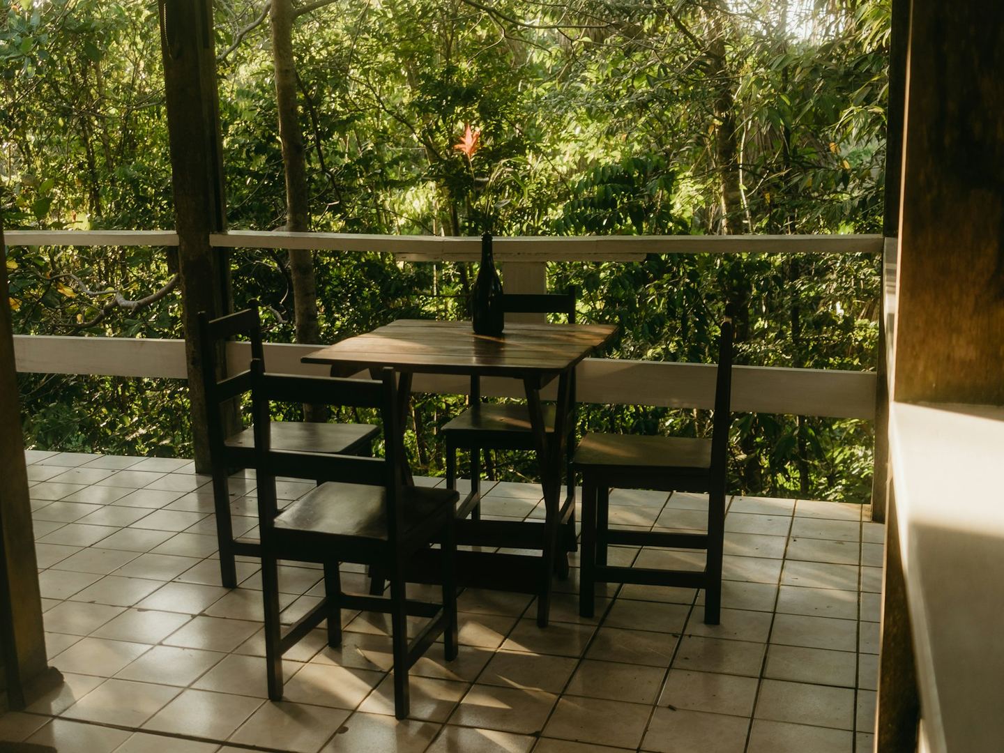 A balcony seen from the doorway, with a wooden table and four chairs on a tiled floor, a white rail and green beyond it.