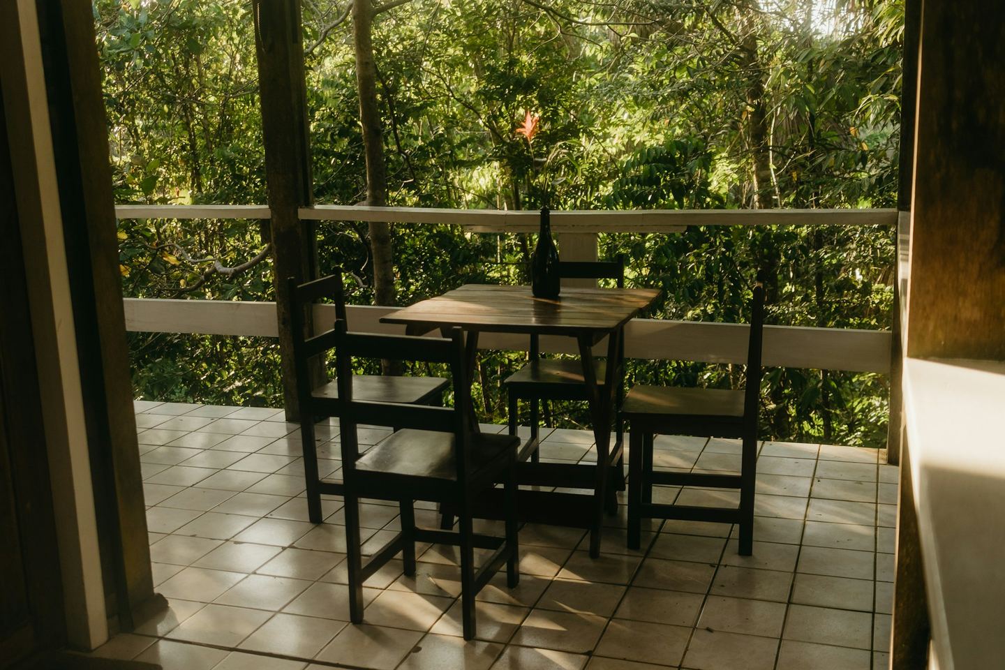 A balcony seen from the doorway, with a wooden table and four chairs on a tiled floor, a white rail and green beyond it.