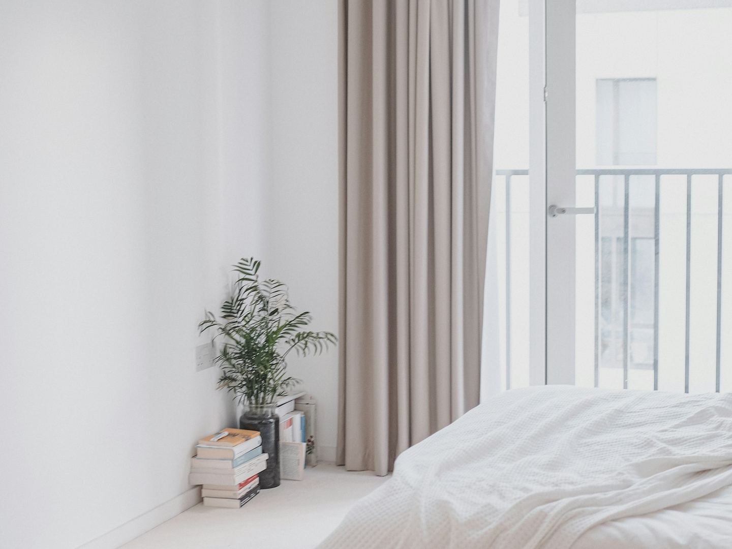 A white bedroom with a linen curtain, a plant on a stack of books and a balcony door.