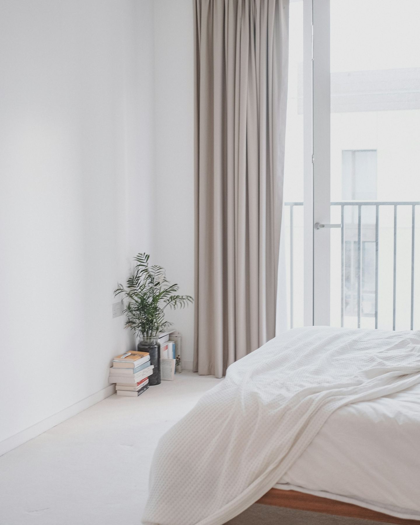 A white bedroom with a linen curtain, a plant on a stack of books and a balcony door.