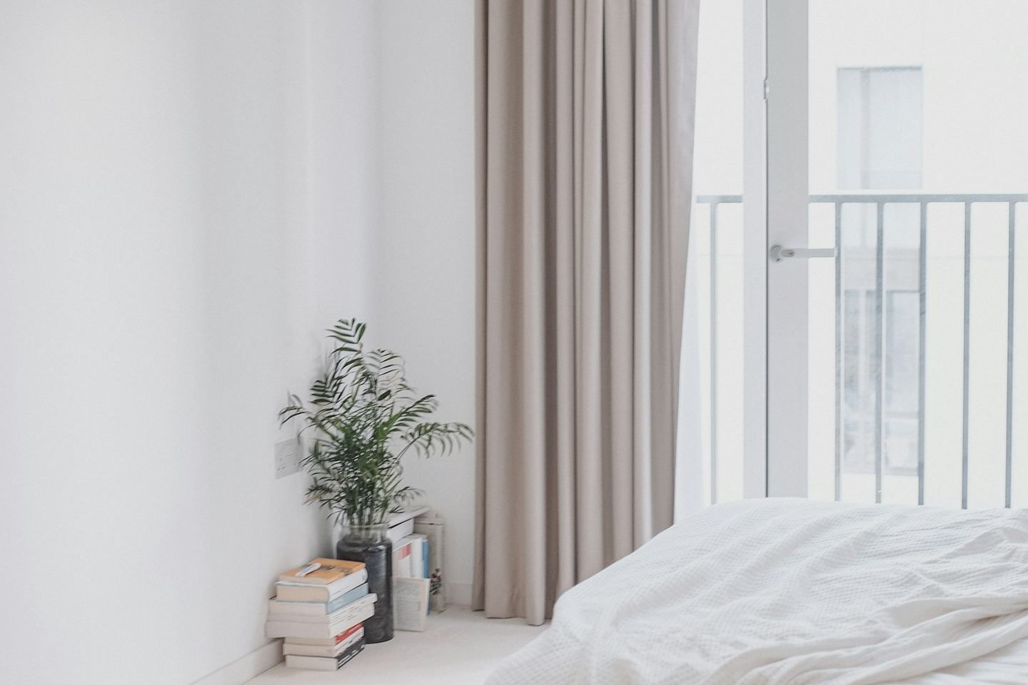 A white bedroom with a linen curtain, a plant on a stack of books and a balcony door.