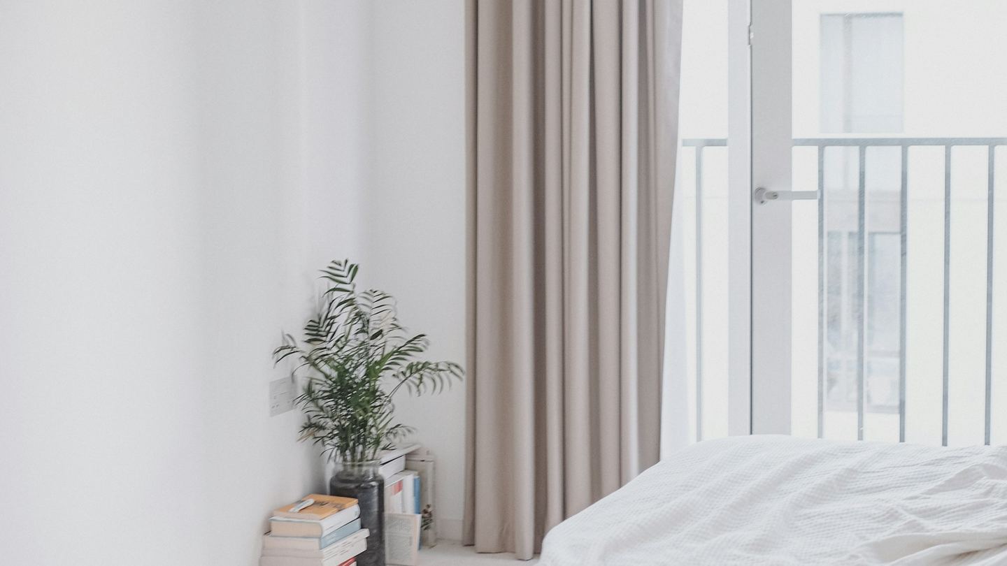 A white bedroom with a linen curtain, a plant on a stack of books and a balcony door.