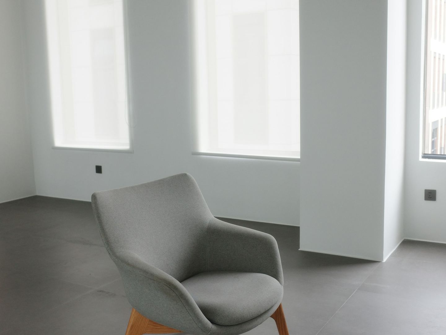 A single grey armchair standing on an open floor in a pale empty room, its back to the windows.