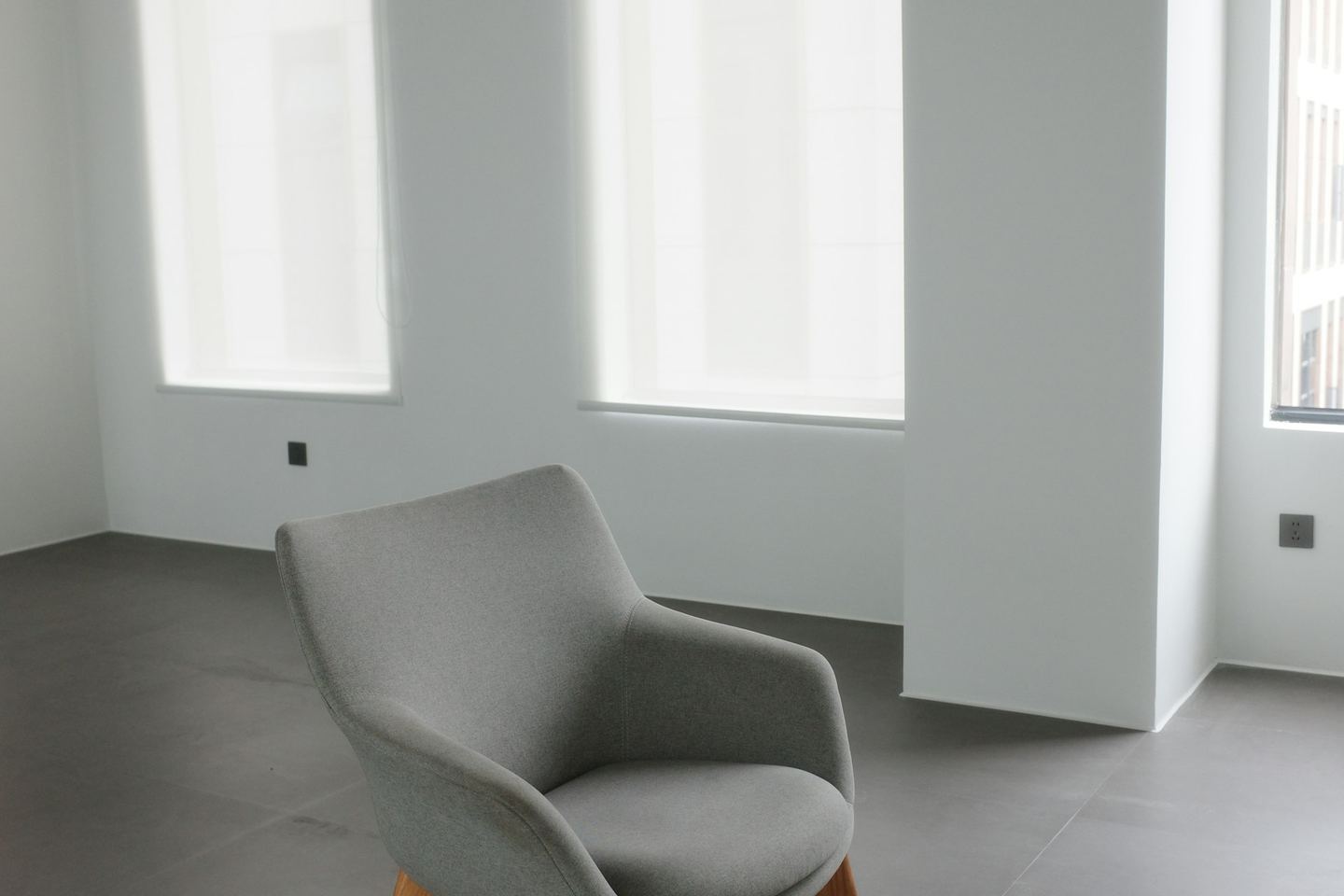 A single grey armchair standing on an open floor in a pale empty room, its back to the windows.