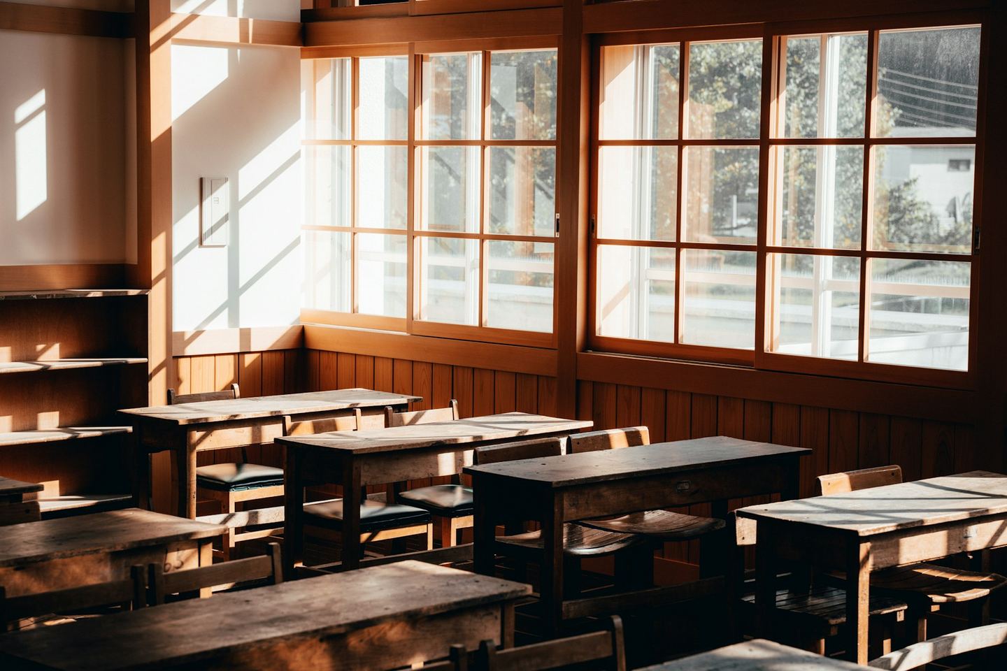 An empty classroom with wooden desks in rows and tall windows along one wall.