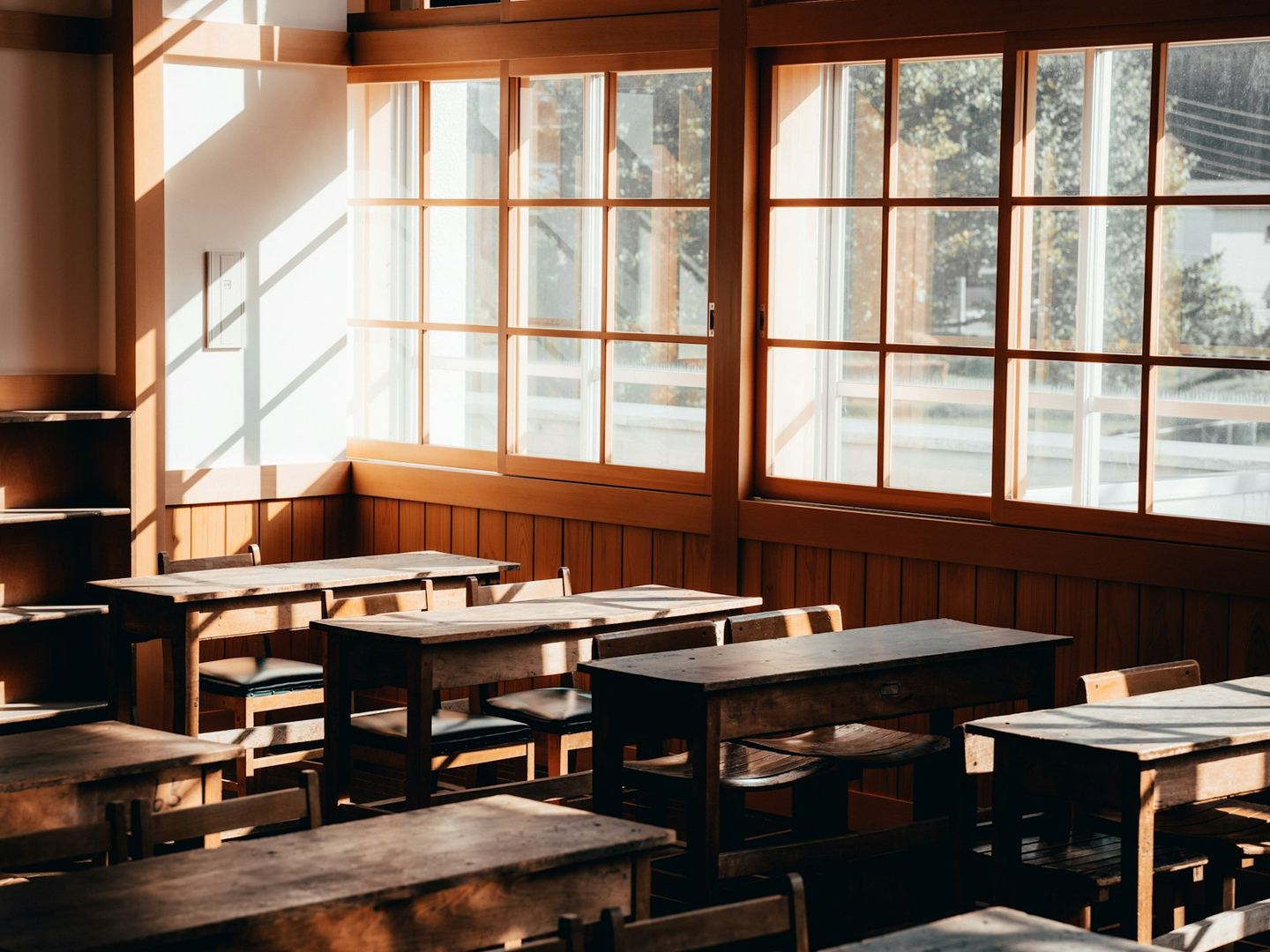 An empty classroom with wooden desks in rows and tall windows along one wall.