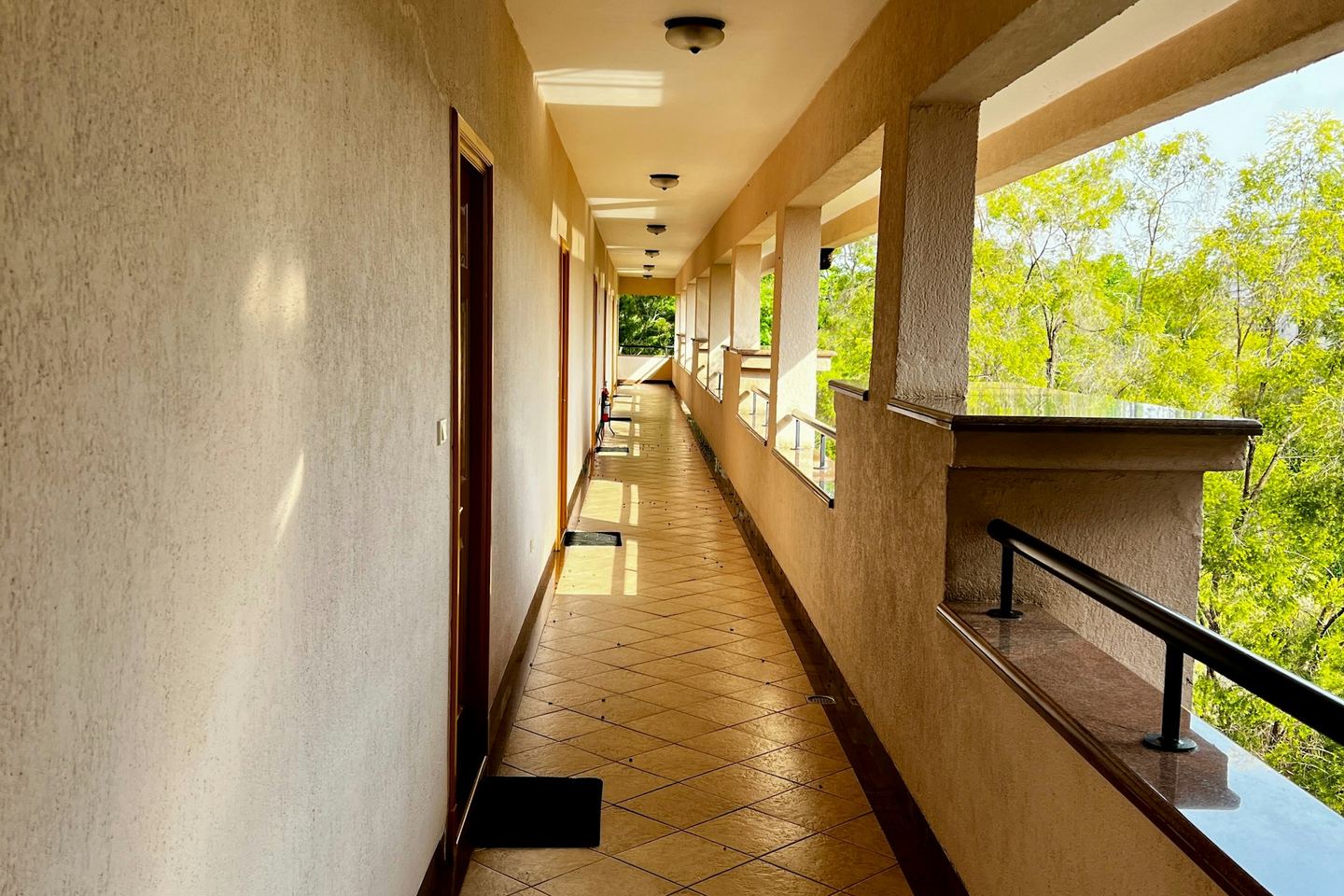 A long, sunlit corridor of an apartment block with doors on one side and the trees beyond the balustrade on the other.