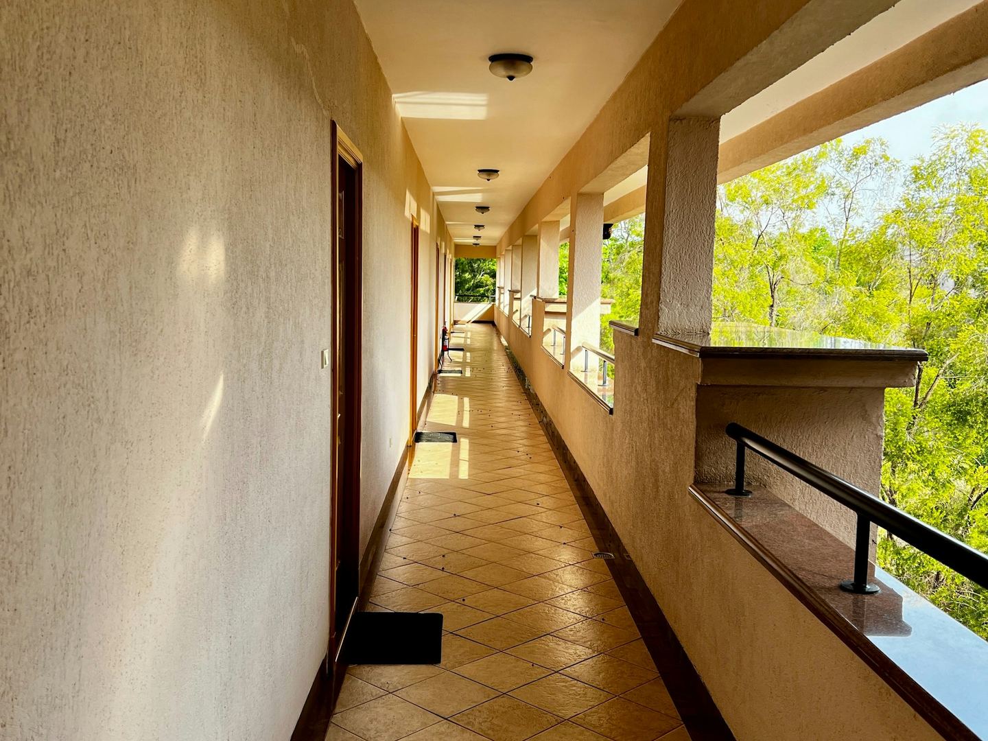 A long, sunlit corridor of an apartment block with doors on one side and the trees beyond the balustrade on the other.