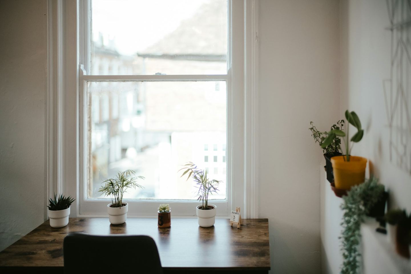 A desk with small plants under a sash window, a chair before it.