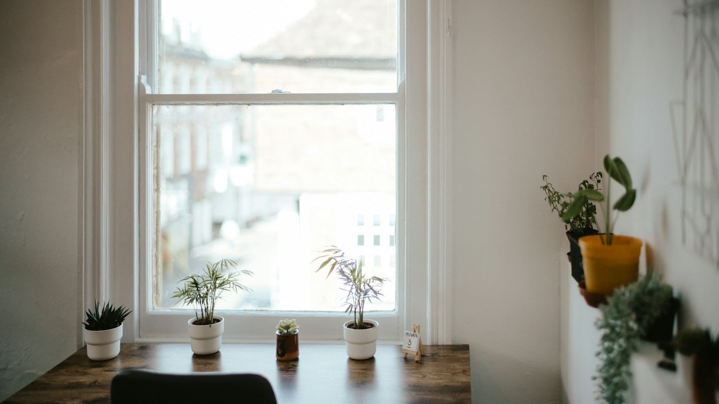 A desk with small plants under a sash window, a chair before it.