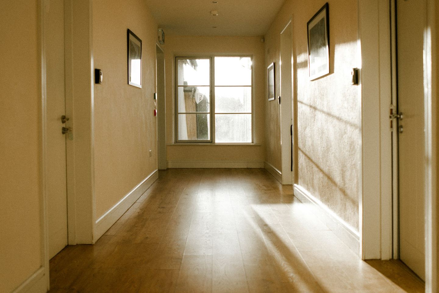 A warm corridor with doors on either side and a window at its end, sun on the floor.