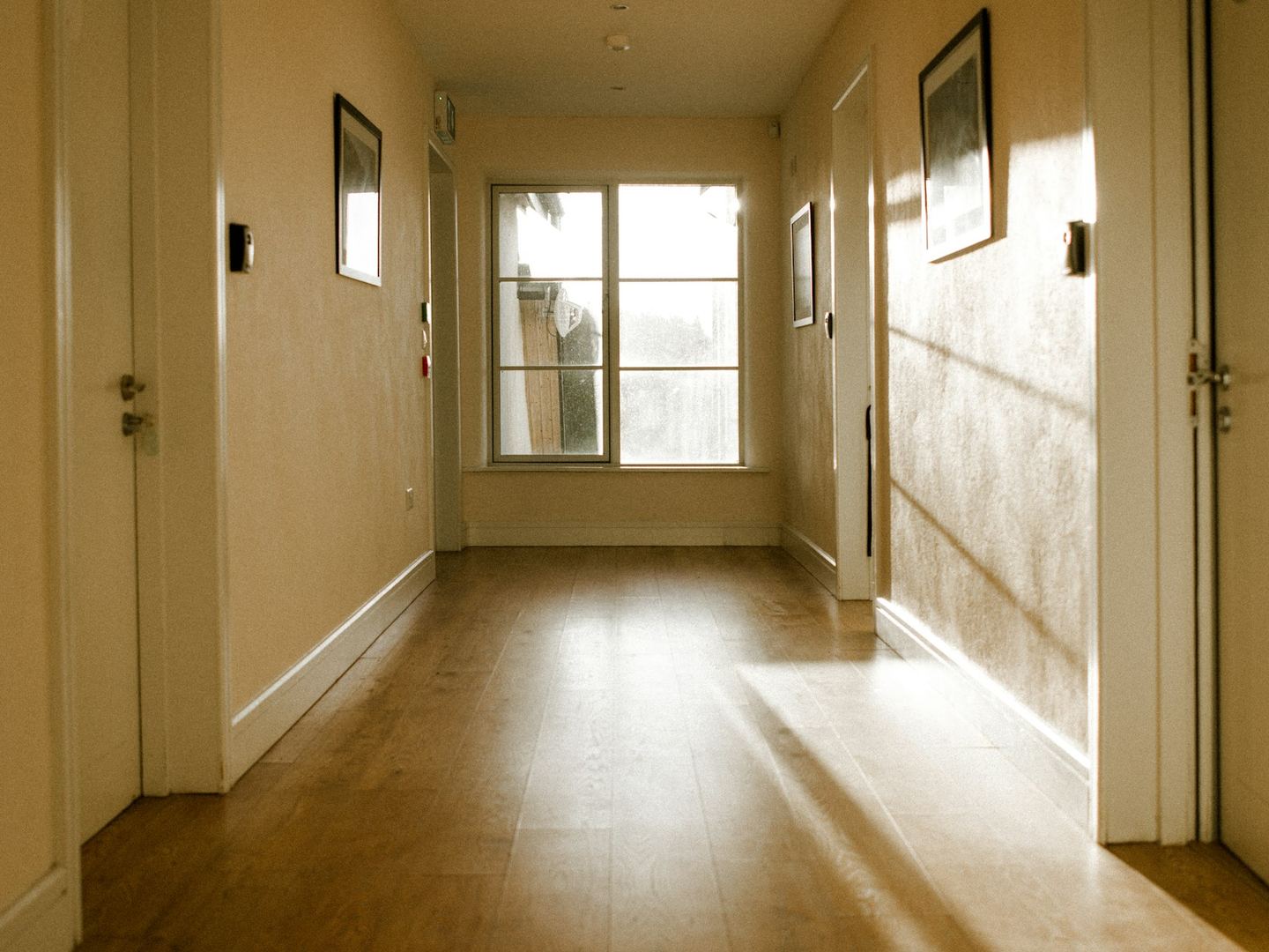 A warm corridor with doors on either side and a window at its end, sun on the floor.