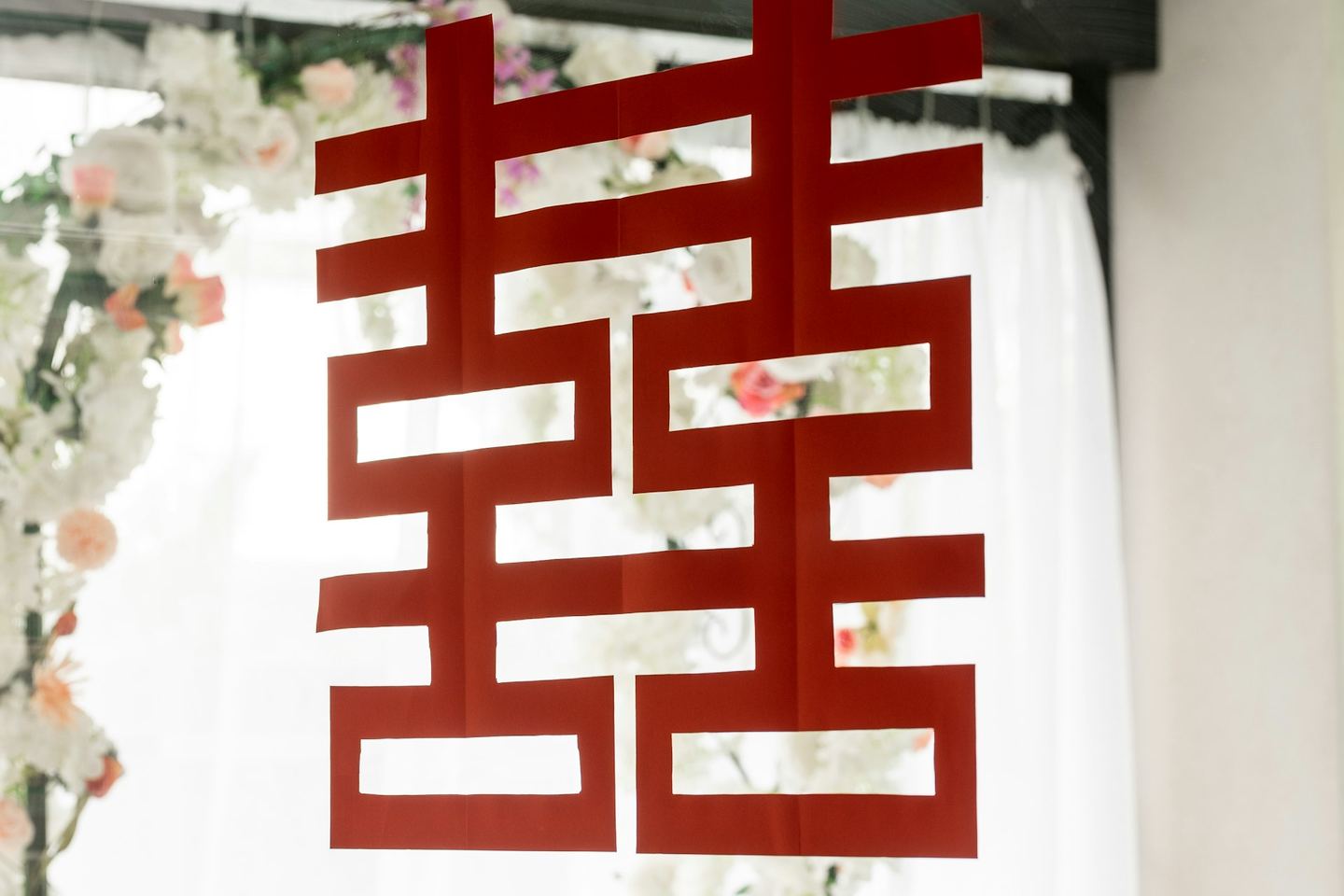The double happiness character cut in red paper, hung in a bright window against white flowers.