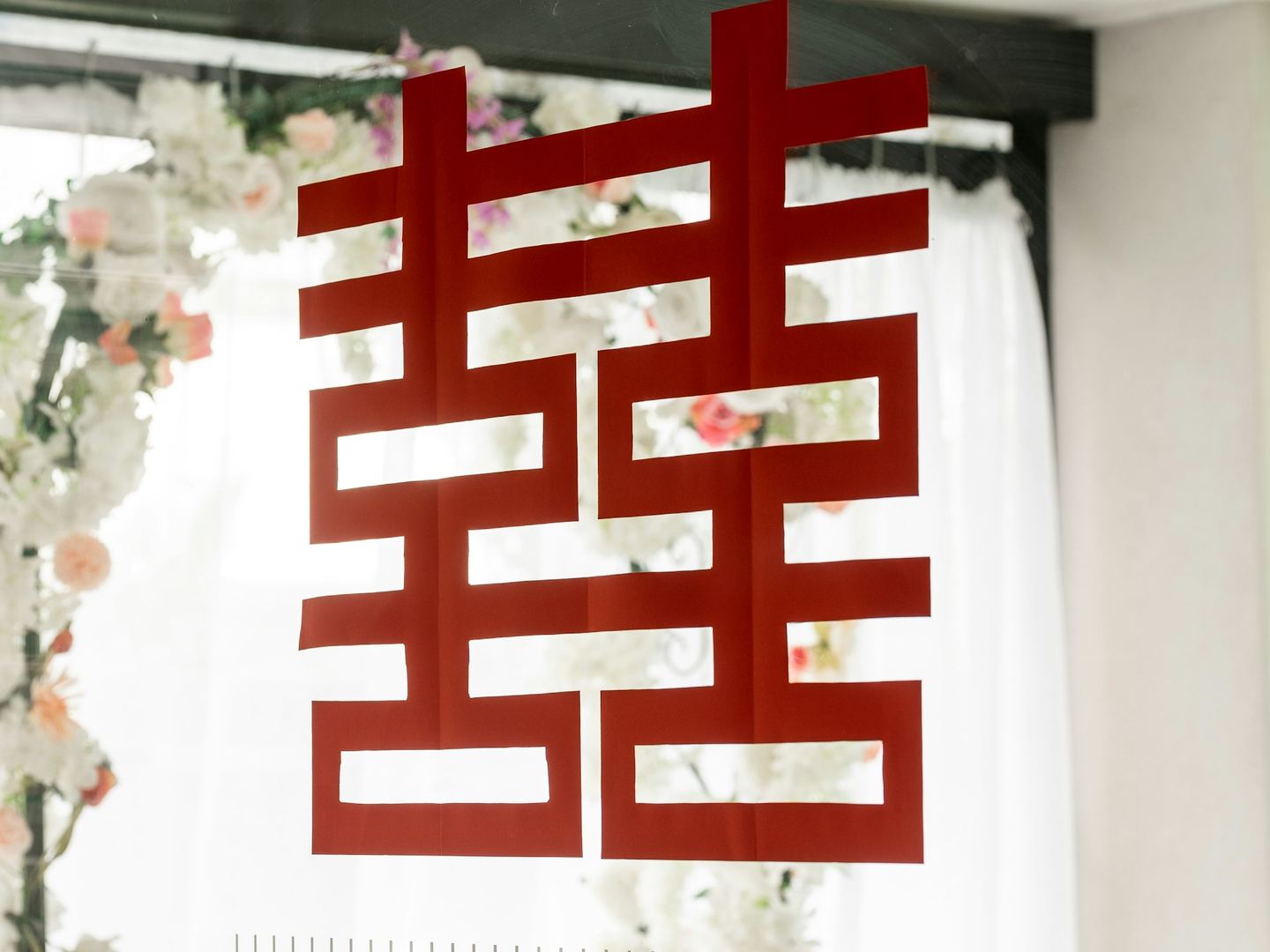 The double happiness character cut in red paper, hung in a bright window against white flowers.
