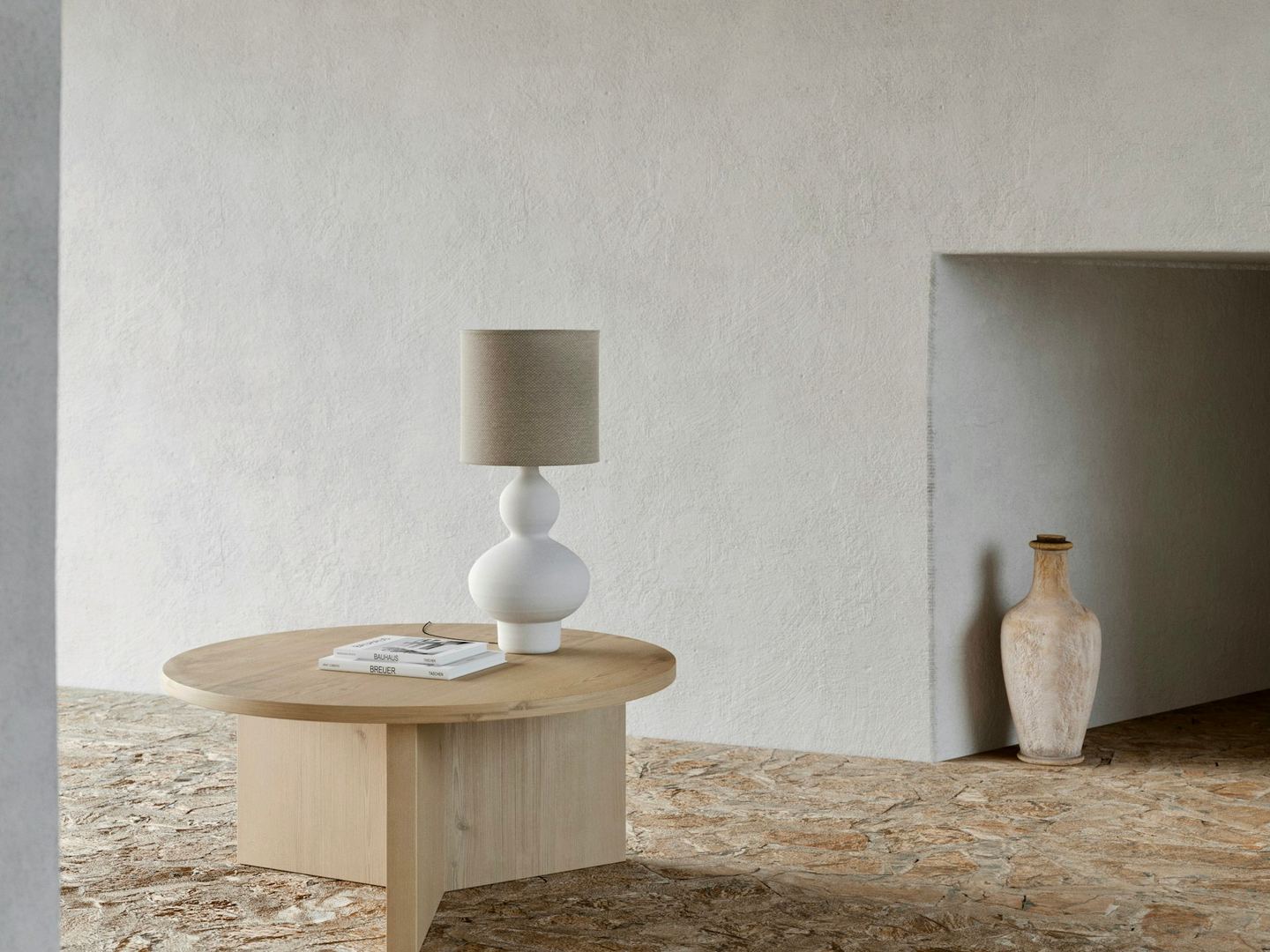 A round wooden table with a ceramic lamp before a white plaster wall, a clay vase in a niche, a stone floor.
