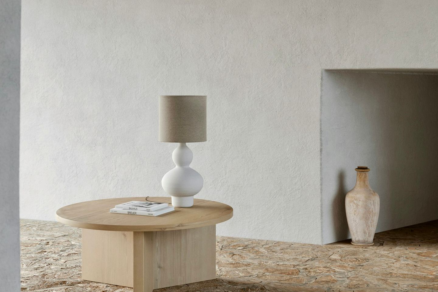 A round wooden table with a ceramic lamp before a white plaster wall, a clay vase in a niche, a stone floor.
