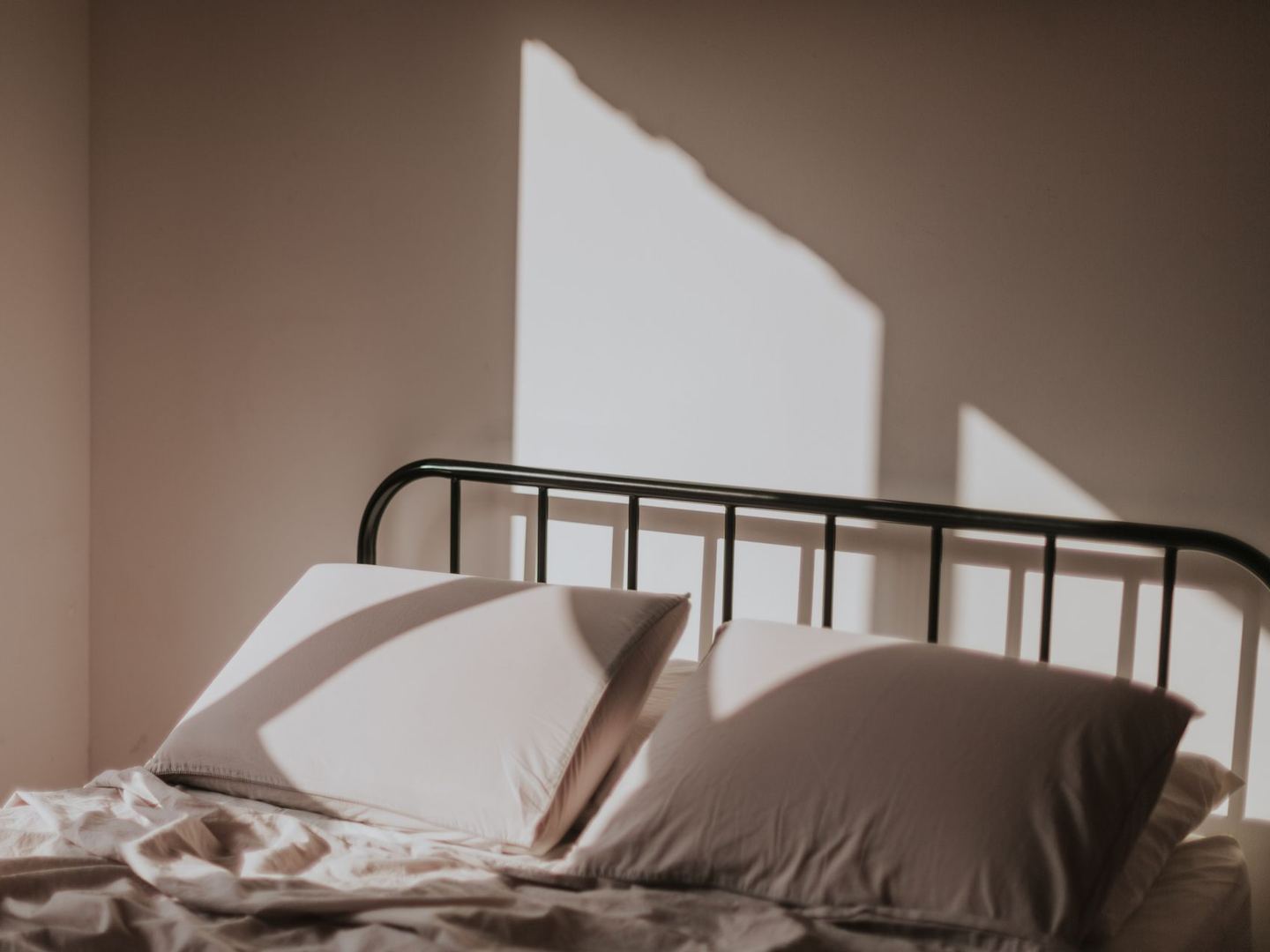 A made bed with a black metal frame against a bare wall, with morning light throwing a hard-edged shadow across the pillows.