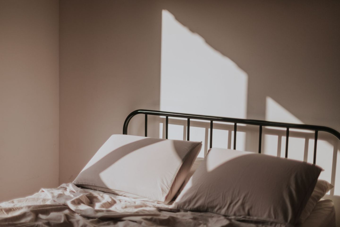 A made bed with a black metal frame against a bare wall, with morning light throwing a hard-edged shadow across the pillows.