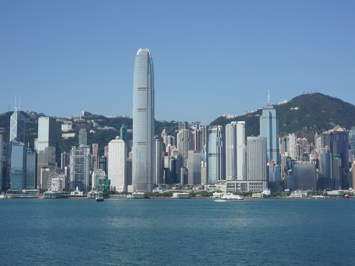 The towers of Central, Hong Kong, on the harbour with the Peak behind them under a clear sky.