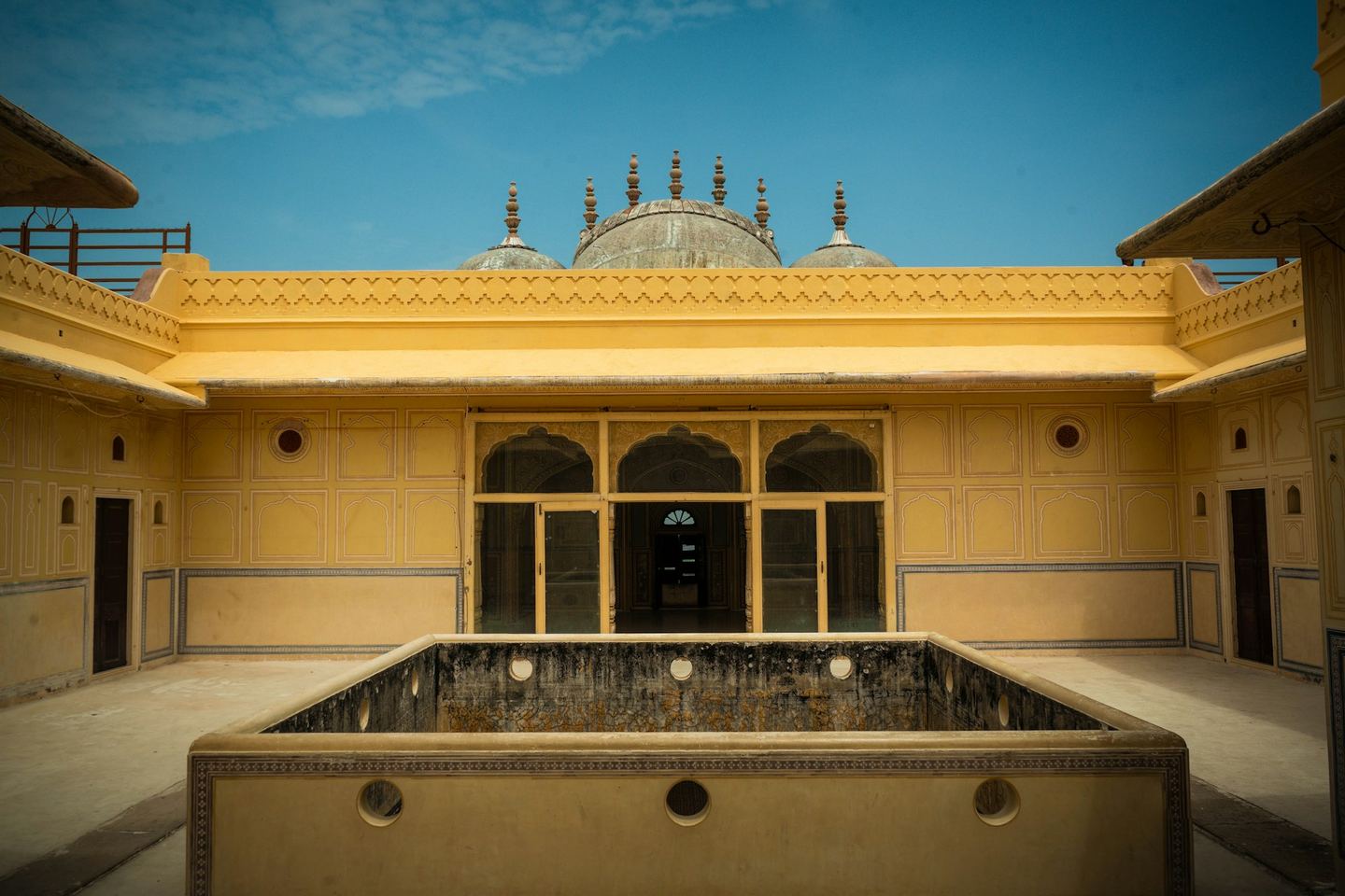 A yellow-walled Rajasthani courtyard with a square water basin at its centre under a blue sky.
