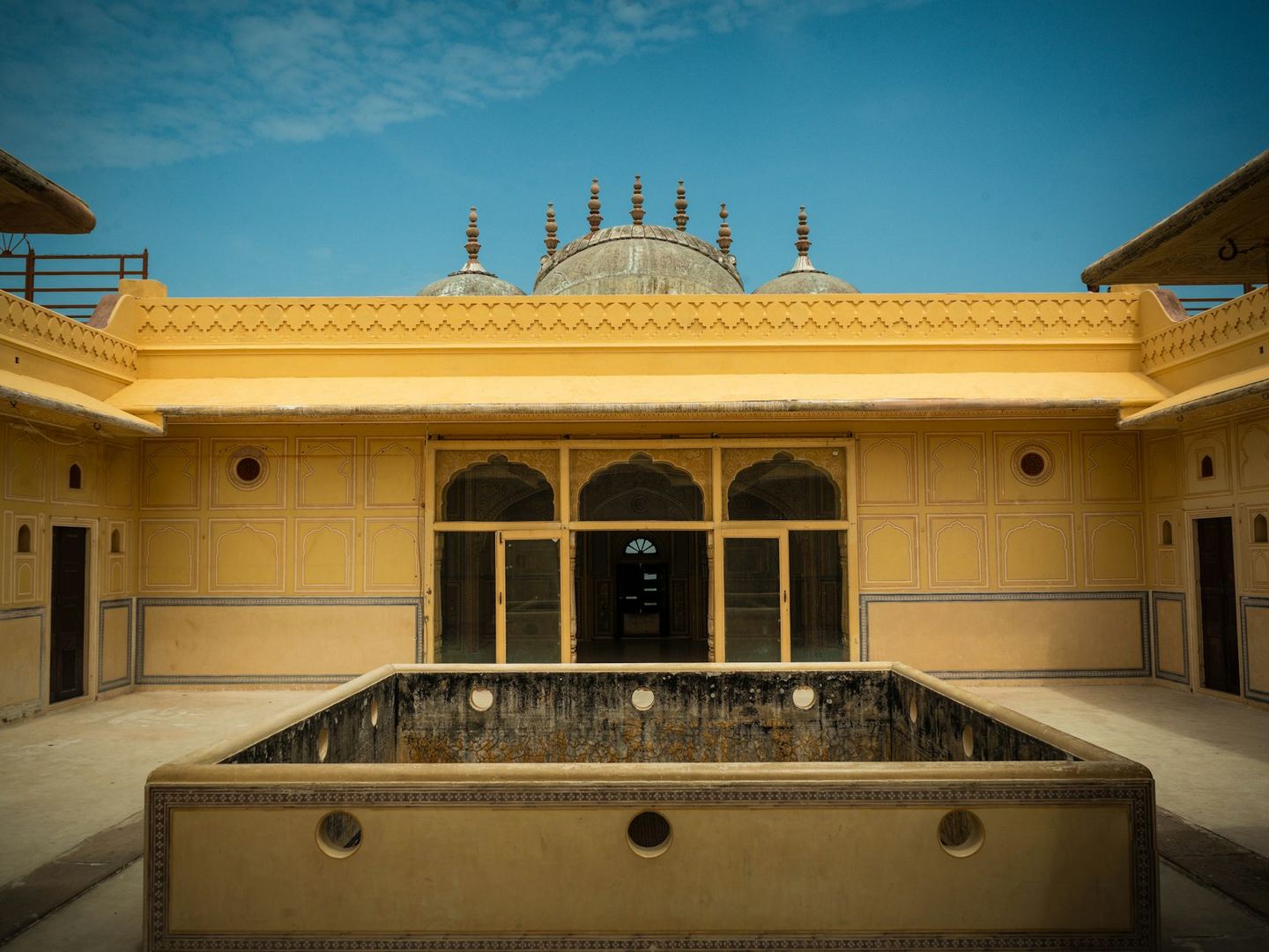 A yellow-walled Rajasthani courtyard with a square water basin at its centre under a blue sky.
