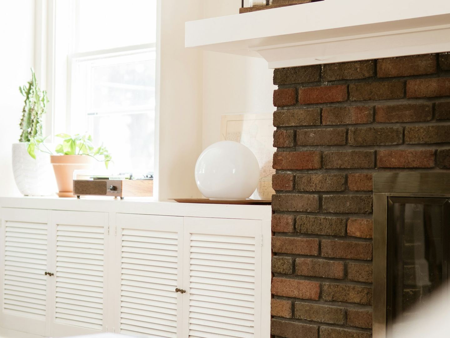 A brick fireplace with a white mantel, candlesticks on it and a sunlit window beside it.