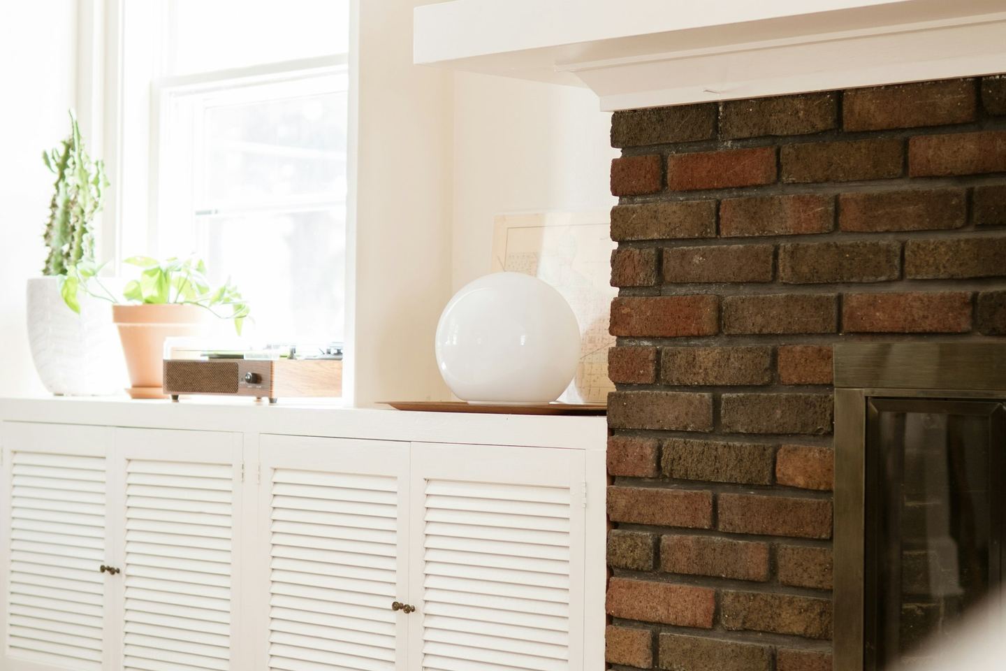 A brick fireplace with a white mantel, candlesticks on it and a sunlit window beside it.