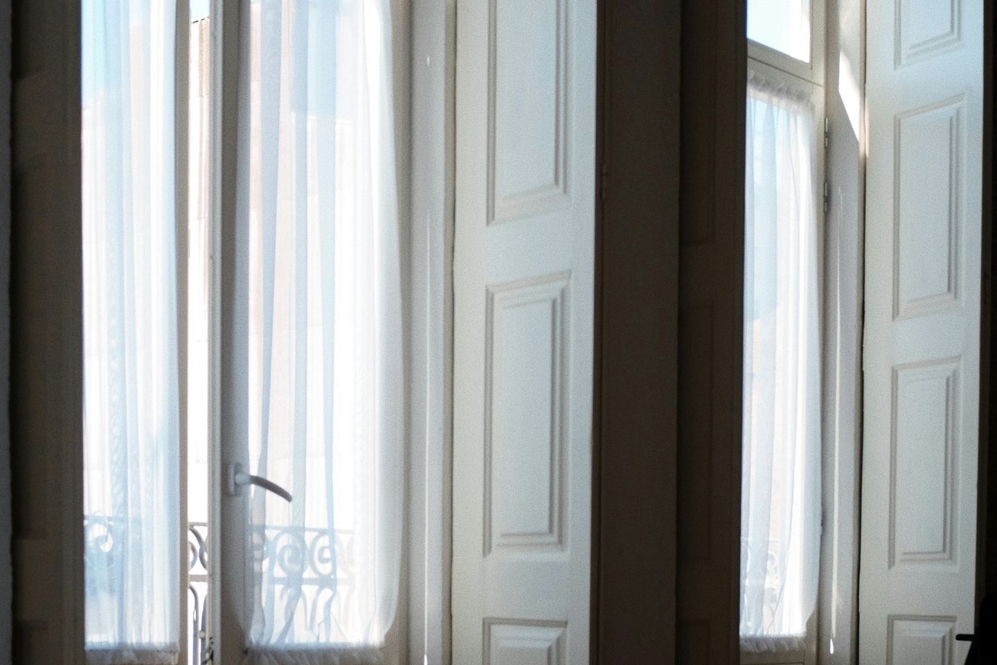 A bed with a white duvet before two tall shuttered windows in morning light.