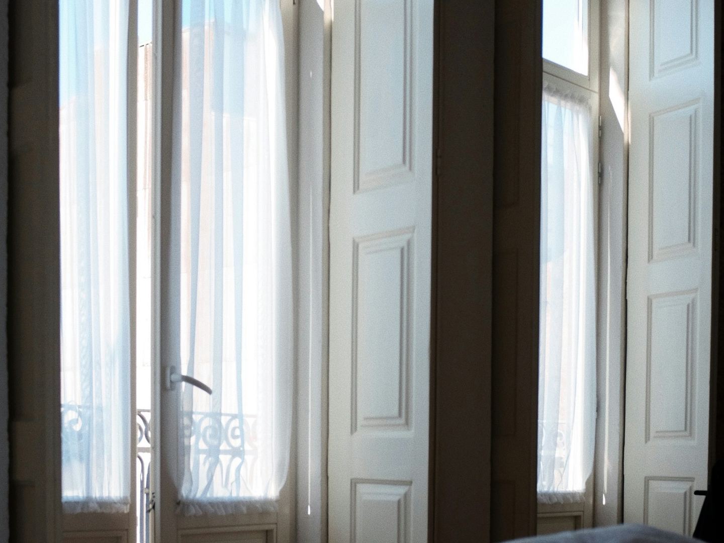A bed with a white duvet before two tall shuttered windows in morning light.