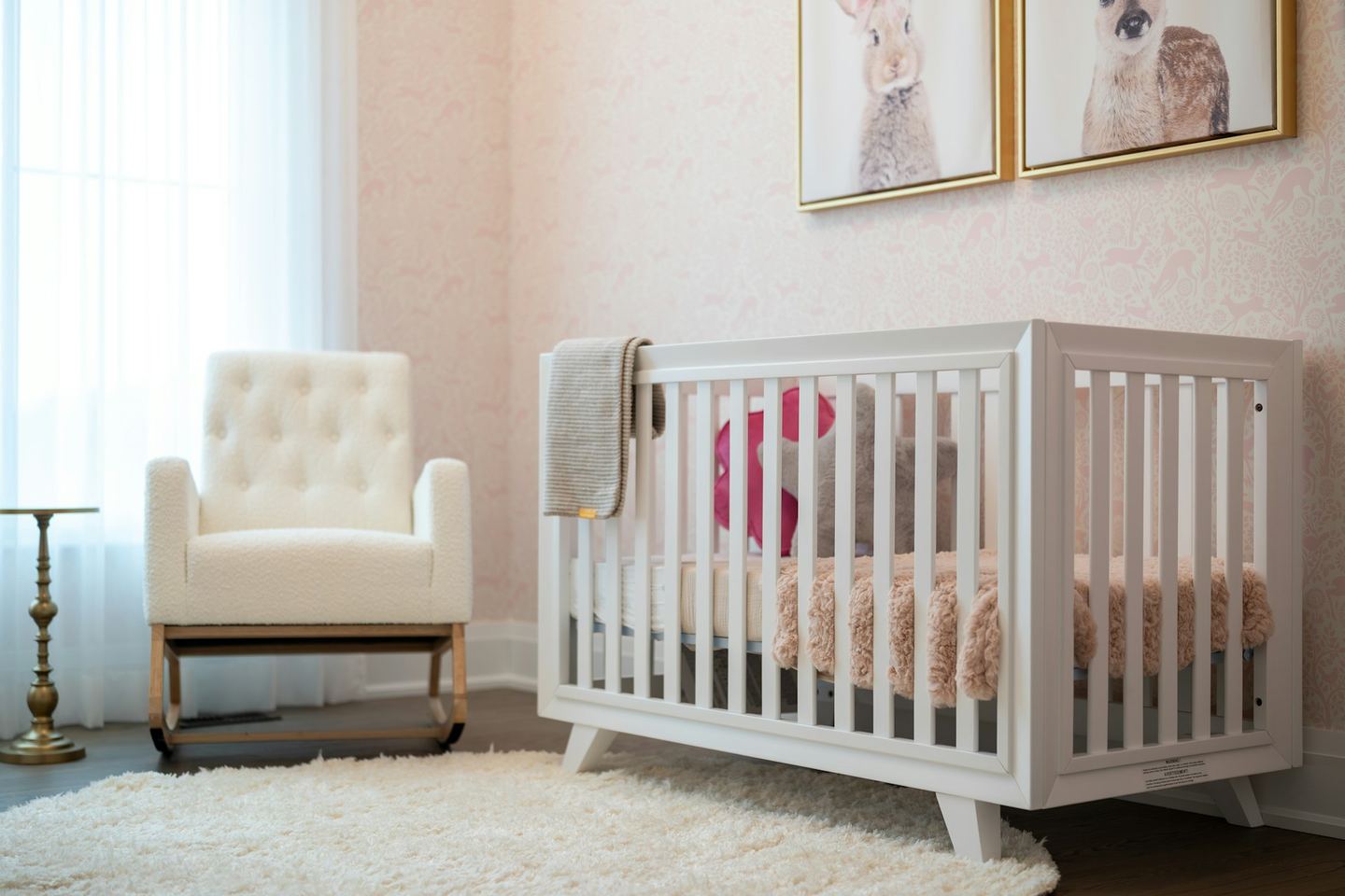 A white cot and a nursing chair in a pale nursery, light through the curtain.