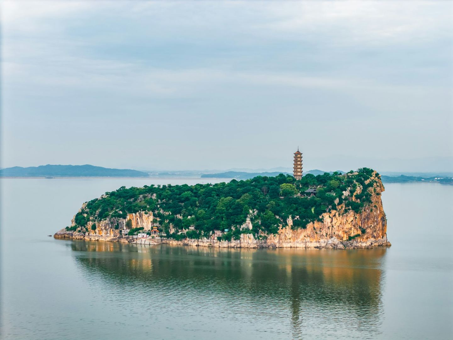 A pagoda on a wooded rocky island in a still lake, hills faint behind.