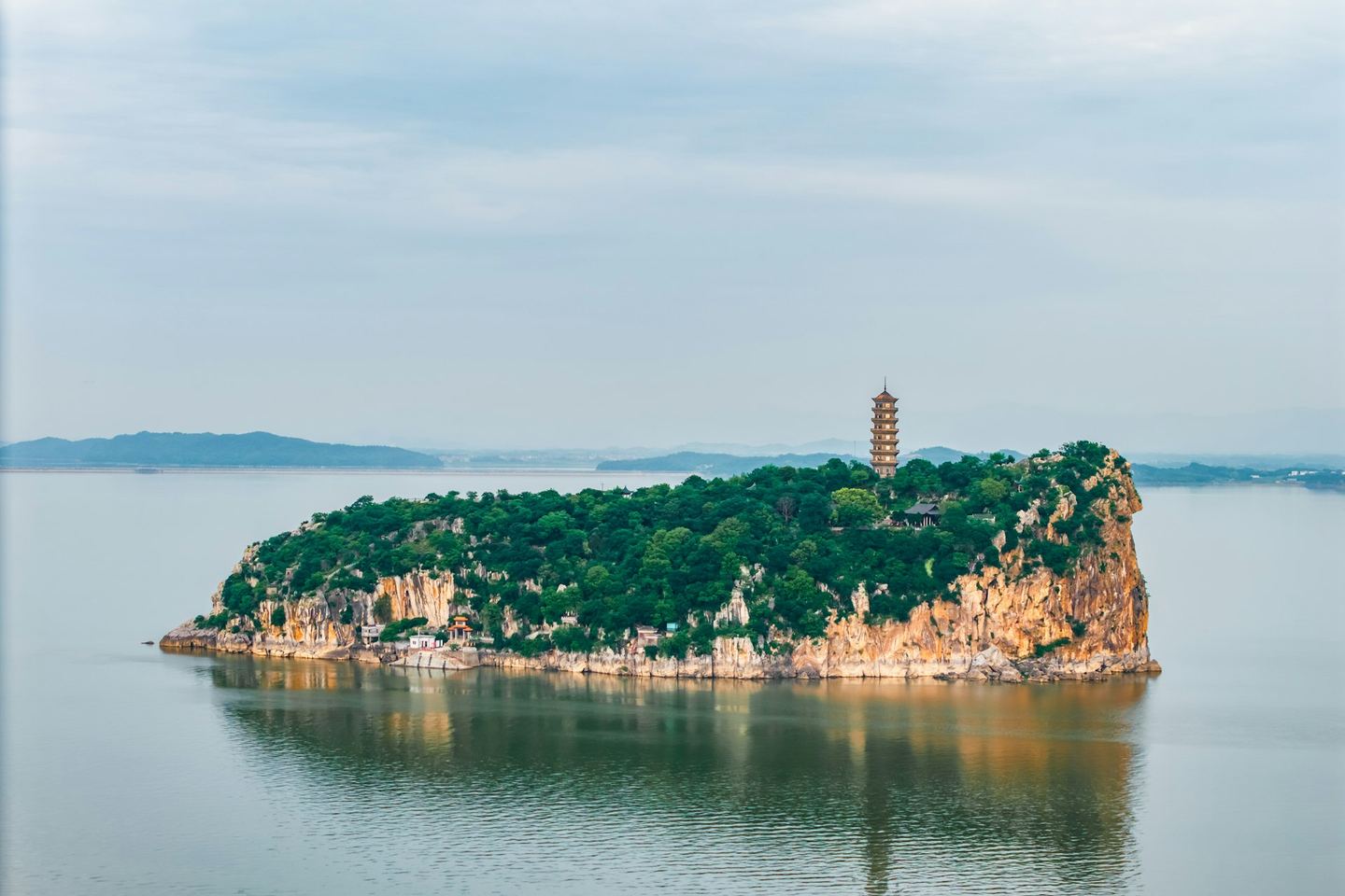 A pagoda on a wooded rocky island in a still lake, hills faint behind.