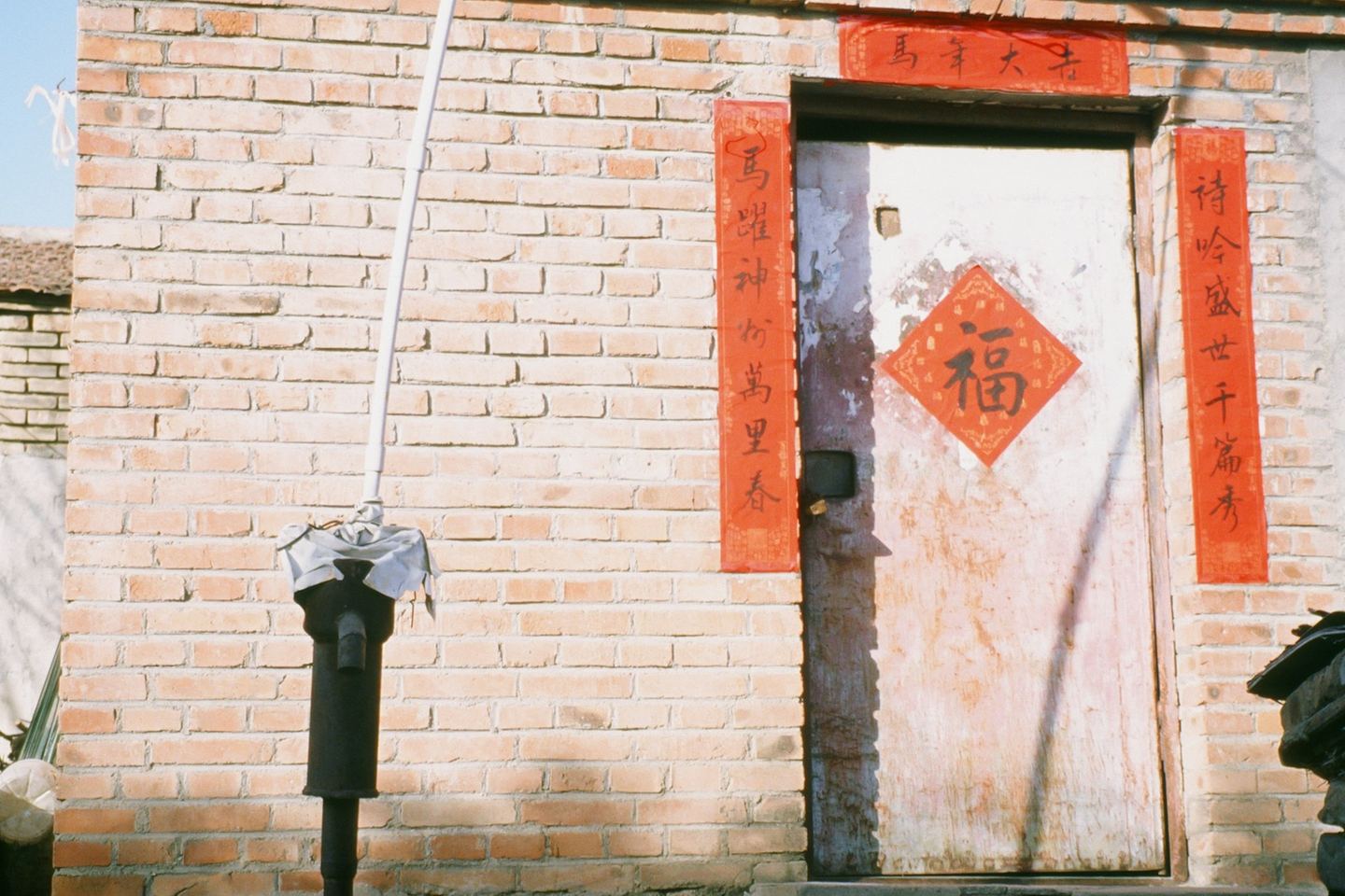 A brick house with red couplets pasted round its door and a fu character on the leaf, in winter sun.