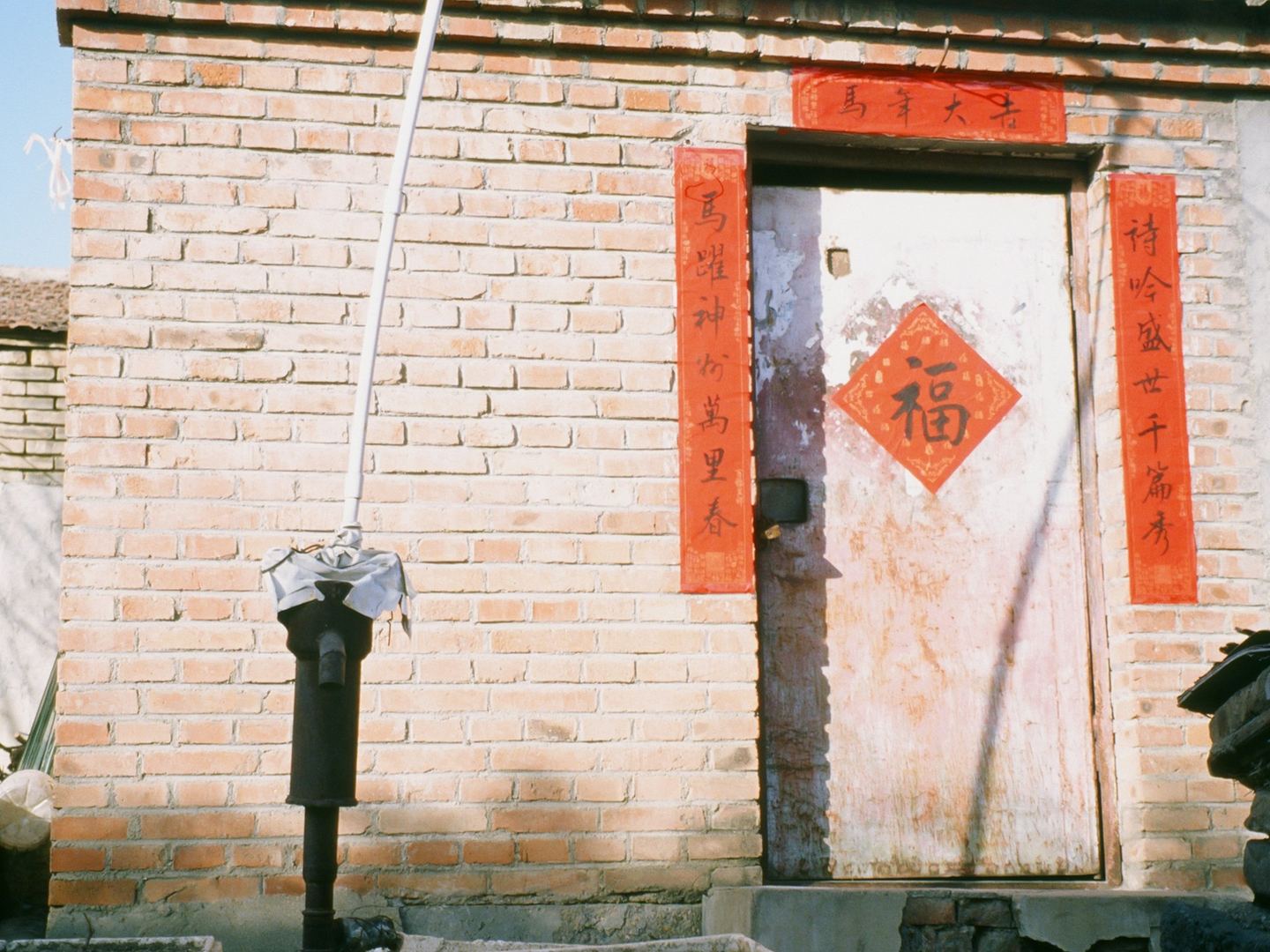A brick house with red couplets pasted round its door and a fu character on the leaf, in winter sun.
