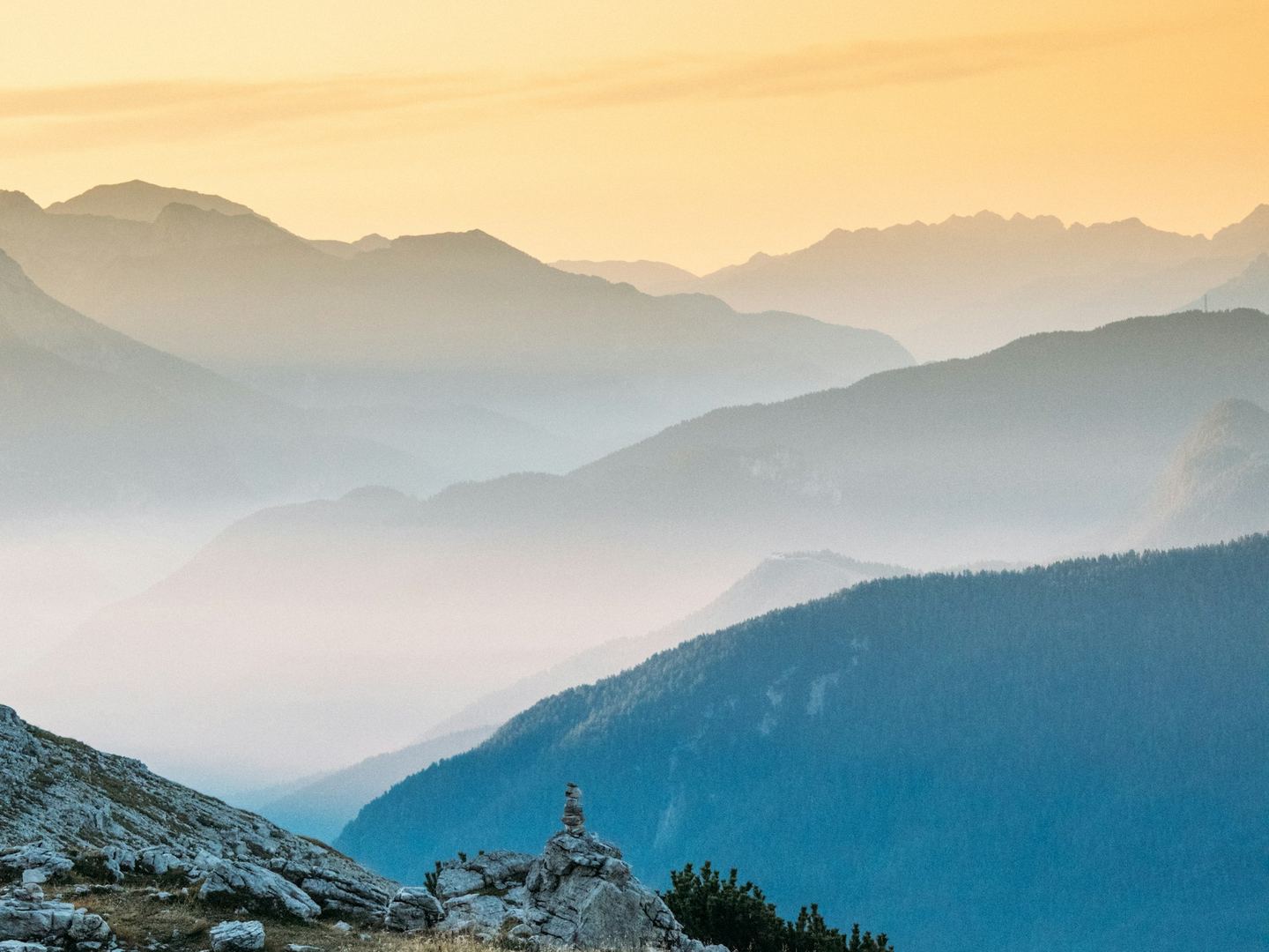 Layered mountain ridges fading into mist under a warm sky, a cairn in the foreground.