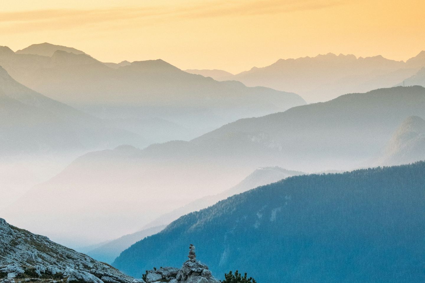 Layered mountain ridges fading into mist under a warm sky, a cairn in the foreground.