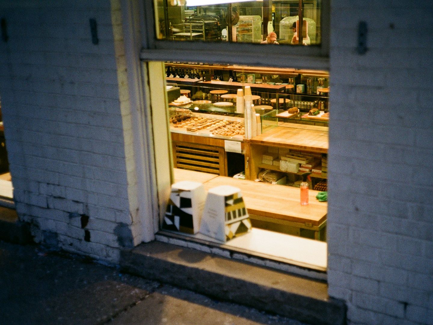 A lit shop seen from the pavement at dusk through its open front: a counter, a glass case of food and shelves along the back wall.
