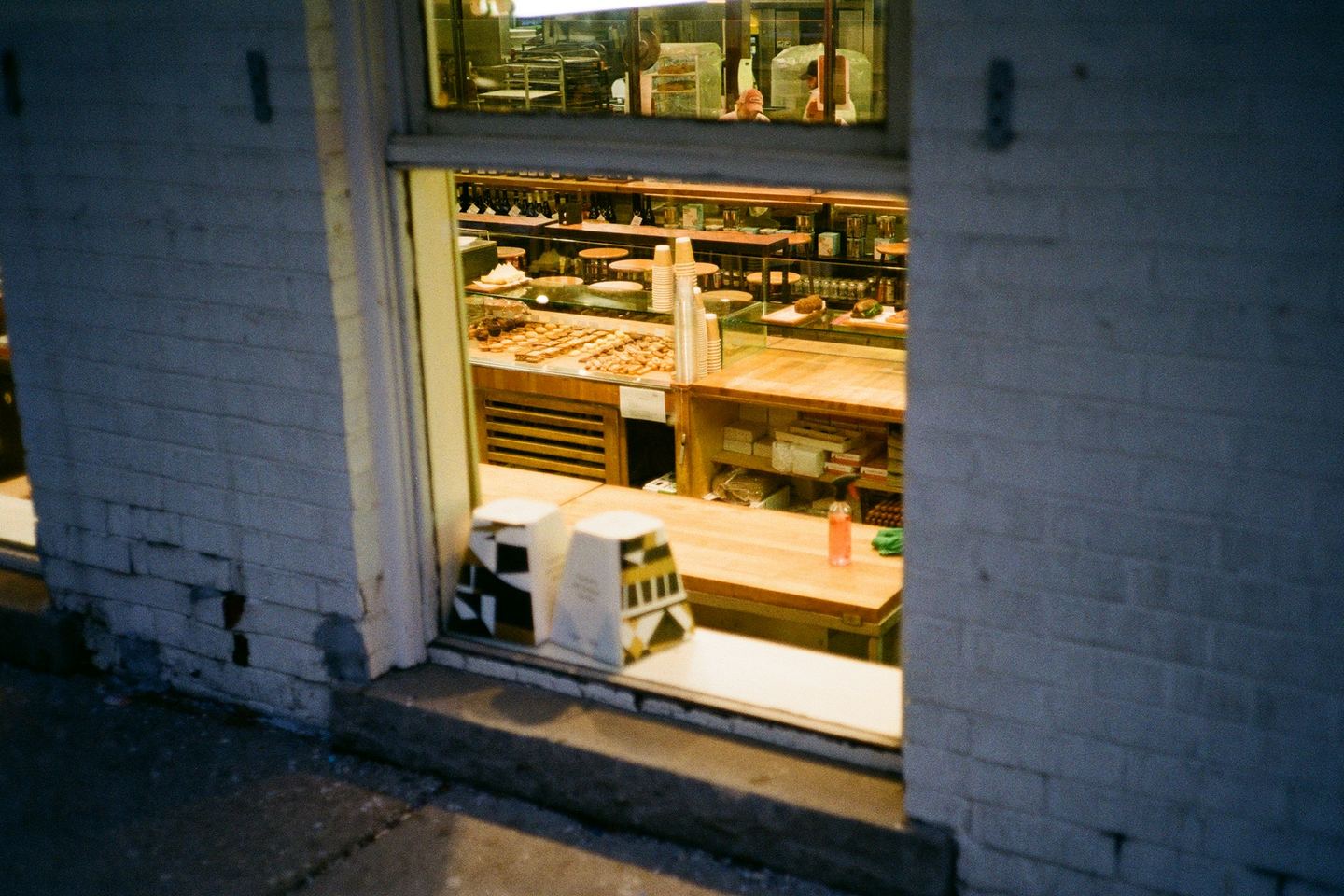 A lit shop seen from the pavement at dusk through its open front: a counter, a glass case of food and shelves along the back wall.