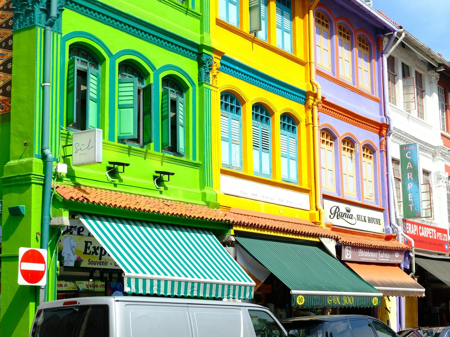 A row of brightly painted shophouses, green, yellow and lilac, with shuttered windows above and awnings over the shops below.