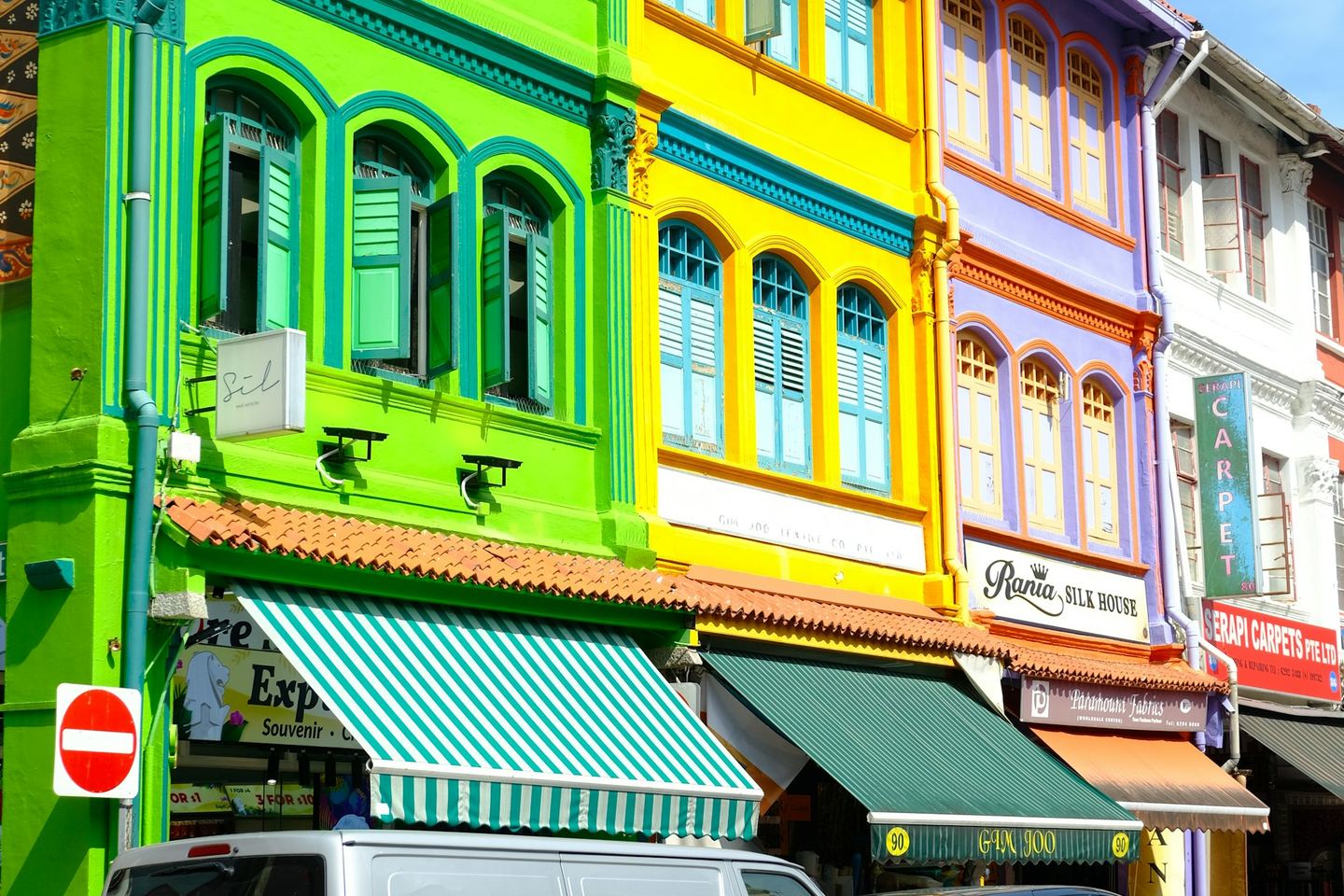 A row of brightly painted shophouses, green, yellow and lilac, with shuttered windows above and awnings over the shops below.