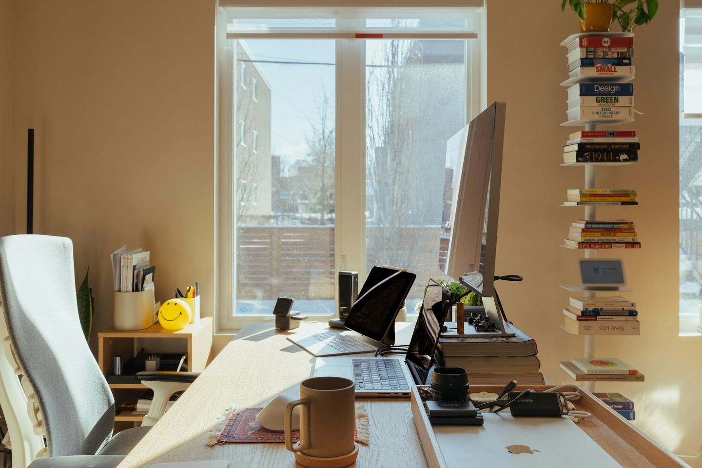 A desk pushed against a sunlit window, a screen and books on it, a wall of shelves at the side.