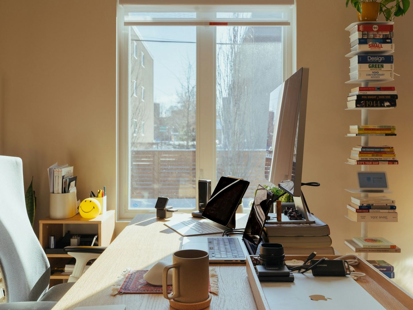 A desk pushed against a sunlit window, a screen and books on it, a wall of shelves at the side.