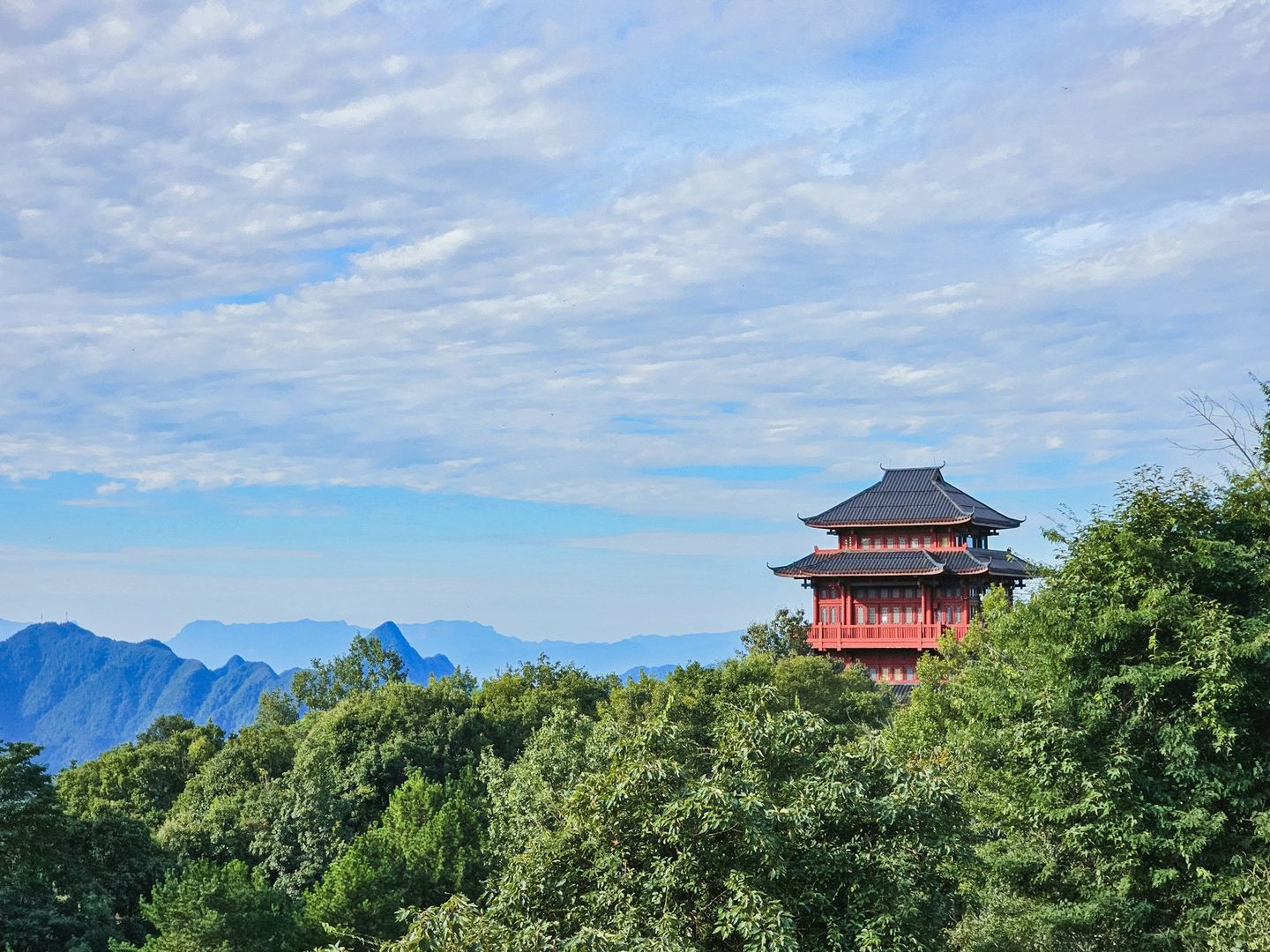 A red two-storey temple pavilion on a wooded hilltop, blue mountain ridges behind under a mackerel sky.