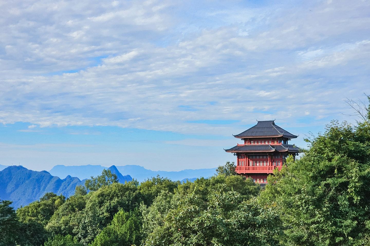 A red two-storey temple pavilion on a wooded hilltop, blue mountain ridges behind under a mackerel sky.